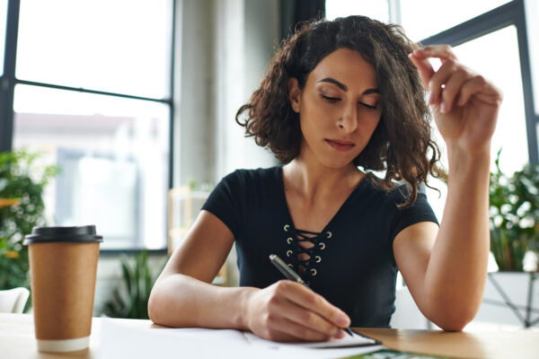 knowledge-sharing, skill-building, concentrated multiracial motivation coach writing in notebook near paper cup with coffee to go, social activism and dedication concept