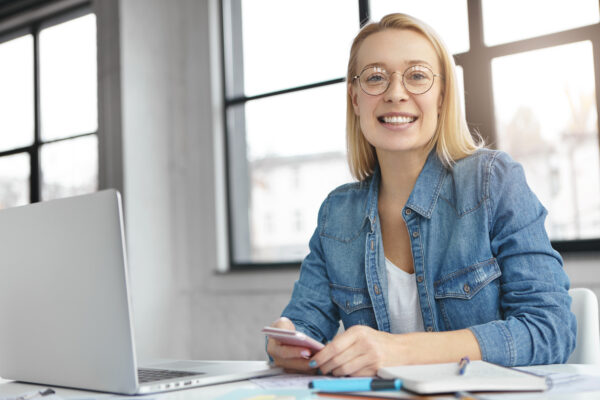 Successful female team leader organizes work of colleagues, inspire them to be active and hard working, uses modern technologies for creating project work, sits at work place over big window