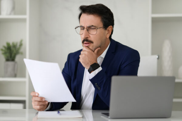 Serious businessman in a blue suit critically examining a document while sitting at his desk with a laptop